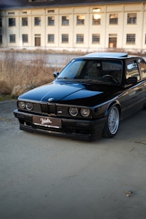 A black vintage BMW car is parked on a concrete surface with a large, beige industrial building in the background. The car is well-maintained, reflecting light on its polished exterior. The scene is set outdoors, with some dry grass and plants visible along the edge of the pavement.