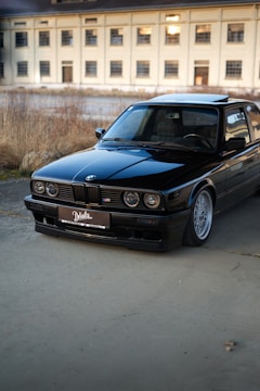 A black vintage BMW car is parked on a concrete surface with a large, beige industrial building in the background. The car is well-maintained, reflecting light on its polished exterior. The scene is set outdoors, with some dry grass and plants visible along the edge of the pavement.