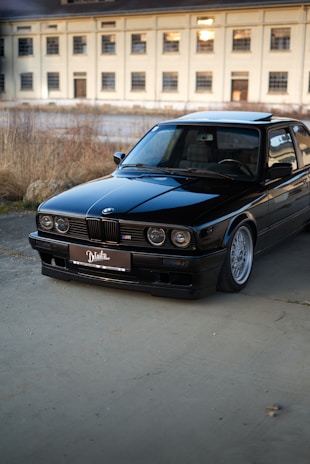 A black vintage BMW car is parked on a concrete surface with a large, beige industrial building in the background. The car is well-maintained, reflecting light on its polished exterior. The scene is set outdoors, with some dry grass and plants visible along the edge of the pavement.