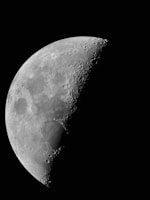 A detailed view of the moon's surface with visible craters and rugged terrain. The moon appears partially illuminated, showcasing its texture against a stark black background.