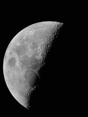 A detailed view of the moon's surface with visible craters and rugged terrain. The moon appears partially illuminated, showcasing its texture against a stark black background.