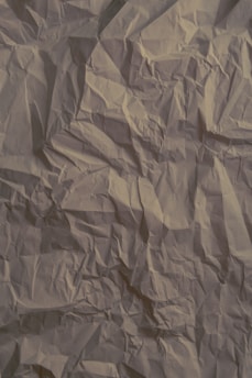 Crinkled beige paper with visible creases and folds throughout the surface, creating a textured and slightly shadowed appearance.