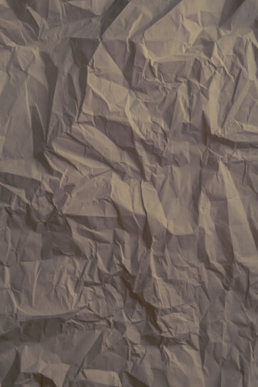 Crinkled beige paper with visible creases and folds throughout the surface, creating a textured and slightly shadowed appearance.