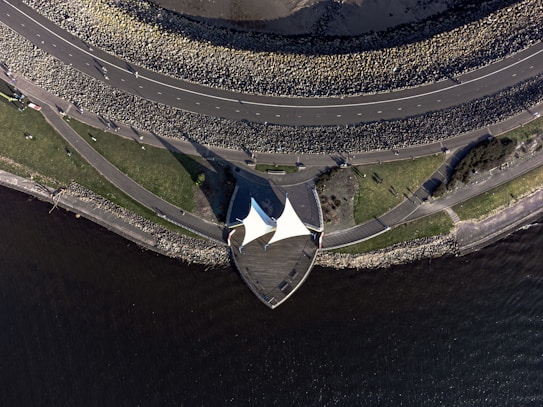 An aerial view of a coastal area featuring a curving road surrounded by large rocks. Below the road, there is a grassy area with pathways and a white tent structure. The shoreline is bordered by water, and some people are visible walking along the paths.
