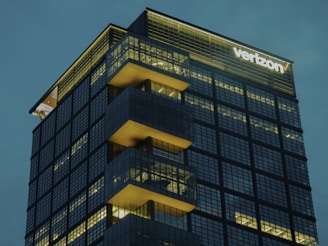 A modern glass skyscraper features illuminated windows and staggered terraces, with a prominent company logo at the top corner glowing against an overcast sky.