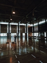 A spacious indoor sports hall features a large wooden floor with white lines marking the area. Several people are sparsely positioned across the floor, including one individual standing in the foreground holding a white cloth. The hall has high ceilings with geometric patterns and large windows that allow natural light to enter, creating a reflective surface on the floor.