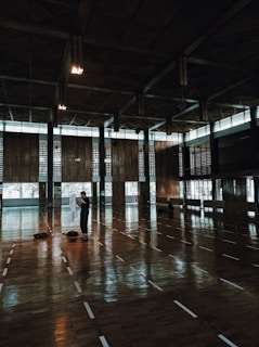 A spacious indoor sports hall features a large wooden floor with white lines marking the area. Several people are sparsely positioned across the floor, including one individual standing in the foreground holding a white cloth. The hall has high ceilings with geometric patterns and large windows that allow natural light to enter, creating a reflective surface on the floor.