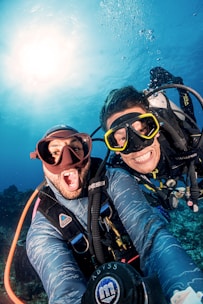 Two scuba divers are submerged underwater, smiling broadly at the camera. They are wearing full diving gear, including wetsuits, masks, and oxygen tanks. Sunlight filters through the clear blue water above them, producing a vibrant and lively atmosphere. Bubbles float around the divers, and the seabed is faintly visible in the background.