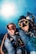 Two scuba divers are submerged underwater, smiling broadly at the camera. They are wearing full diving gear, including wetsuits, masks, and oxygen tanks. Sunlight filters through the clear blue water above them, producing a vibrant and lively atmosphere. Bubbles float around the divers, and the seabed is faintly visible in the background.