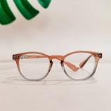 A pair of glasses with a light brown and transparent gradient frame is placed on a smooth, light-colored surface. In the background, part of a green leaf is visible, adding a touch of natural contrast.