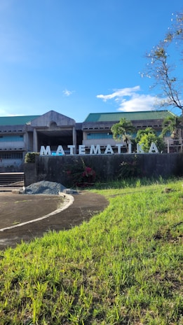 A modern building with a large sign spelling 'MATEMATIKA' is situated behind a grassy area under a bright blue sky. The structure has a concrete facade and is surrounded by greenery, including trees and shrubs.