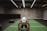 A person in athletic wear is pushing a sled on green turf inside a dimly lit gym with 'Global Fitness' written on the wall. The setting includes exercise equipment and industrial elements, such as visible metal beams and a rolled-up garage door.