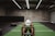 A person in athletic wear is pushing a sled on green turf inside a dimly lit gym with 'Global Fitness' written on the wall. The setting includes exercise equipment and industrial elements, such as visible metal beams and a rolled-up garage door.