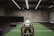 A person in athletic wear is pushing a sled on green turf inside a dimly lit gym with 'Global Fitness' written on the wall. The setting includes exercise equipment and industrial elements, such as visible metal beams and a rolled-up garage door.