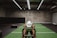 A person in athletic wear is pushing a sled on green turf inside a dimly lit gym with 'Global Fitness' written on the wall. The setting includes exercise equipment and industrial elements, such as visible metal beams and a rolled-up garage door.