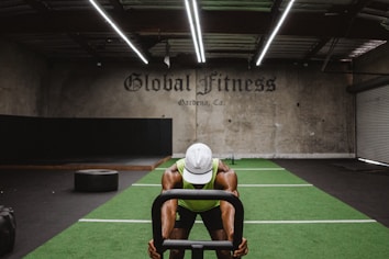 A person in athletic wear is pushing a sled on green turf inside a dimly lit gym with 'Global Fitness' written on the wall. The setting includes exercise equipment and industrial elements, such as visible metal beams and a rolled-up garage door.