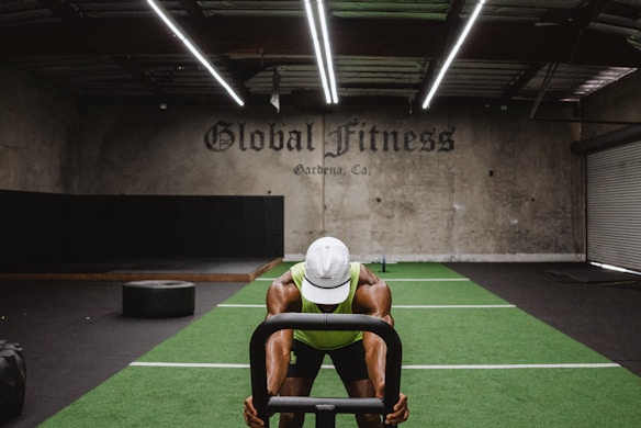 A person in athletic wear is pushing a sled on green turf inside a dimly lit gym with 'Global Fitness' written on the wall. The setting includes exercise equipment and industrial elements, such as visible metal beams and a rolled-up garage door.