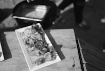 A black and white image featuring a wooden table with a rectangular white plate holding a meal. The food appears to be a combination of vegetables and a grilled dish, possibly fish or chicken. A pot can be seen in the background, and part of a person's hand is visible near the top edge.