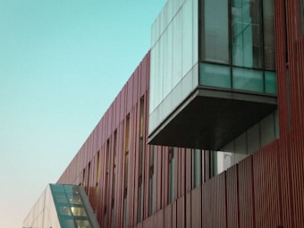 A modern architectural structure with a combination of red vertical panels and large glass windows. The building features an angular glass facade and a prominent overhanging section, creating a dynamic and contemporary design.