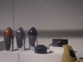 The scene displays a collection of retro-style household or kitchen items set on a neutral surface under soft lighting. There are three metallic containers with colored tops in shades of brown, silver, and black. Additionally, a vintage radio and a light-colored ceramic kettle are also visible.