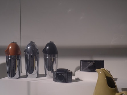The scene displays a collection of retro-style household or kitchen items set on a neutral surface under soft lighting. There are three metallic containers with colored tops in shades of brown, silver, and black. Additionally, a vintage radio and a light-colored ceramic kettle are also visible.