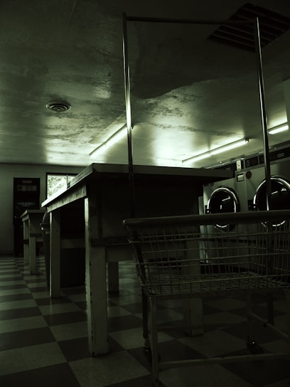 A dimly lit laundromat with industrial-style washing machines lining the wall. The floors are checkered and there are long, metal tables and a laundry cart in the foreground. Fluorescent lights cast a green hue over the scene, with minimal natural light coming from a small window.