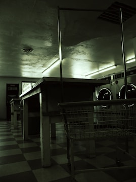 A dimly lit laundromat with industrial-style washing machines lining the wall. The floors are checkered and there are long, metal tables and a laundry cart in the foreground. Fluorescent lights cast a green hue over the scene, with minimal natural light coming from a small window.
