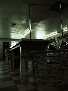 A dimly lit laundromat with industrial-style washing machines lining the wall. The floors are checkered and there are long, metal tables and a laundry cart in the foreground. Fluorescent lights cast a green hue over the scene, with minimal natural light coming from a small window.