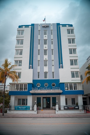 A multi-story Art Deco style hotel featuring a combination of white and blue colors. The facade includes vertical lines and geometric design elements. In front there are palm trees and the entrance is decorated with small plants.