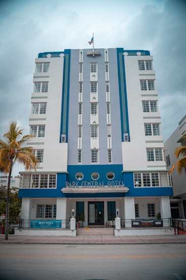 A multi-story Art Deco style hotel featuring a combination of white and blue colors. The facade includes vertical lines and geometric design elements. In front there are palm trees and the entrance is decorated with small plants.
