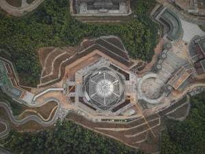 An aerial view of a symmetrical, geometric structure under construction, surrounded by a mix of greenery and other buildings. The central building is shaped like a hexagon with clear sections and scaffolding or cranes visible, indicating ongoing construction. The surrounding landscape includes winding roads and green forested areas.