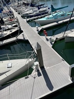 A marina filled with numerous boats docked along wooden piers. Several sailboats with masts and rigging are neatly aligned in greenish water. The scene includes dock accessories such as a life preserver and bollards.