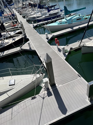 A marina filled with numerous boats docked along wooden piers. Several sailboats with masts and rigging are neatly aligned in greenish water. The scene includes dock accessories such as a life preserver and bollards.