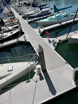 A marina filled with numerous boats docked along wooden piers. Several sailboats with masts and rigging are neatly aligned in greenish water. The scene includes dock accessories such as a life preserver and bollards.