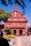 A red brick colonial-style church with a white cross, surrounded by well-maintained topiary and flower gardens. A clear blue sky frames the structure, with people walking nearby, some using umbrellas for shade. Motorbikes are parked on the pathway leading to the church entrance.