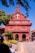 A red brick colonial-style church with a white cross, surrounded by well-maintained topiary and flower gardens. A clear blue sky frames the structure, with people walking nearby, some using umbrellas for shade. Motorbikes are parked on the pathway leading to the church entrance.