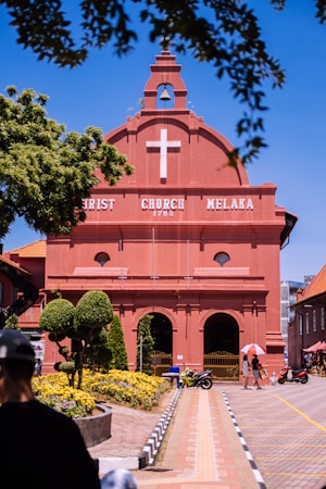 A red brick colonial-style church with a white cross, surrounded by well-maintained topiary and flower gardens. A clear blue sky frames the structure, with people walking nearby, some using umbrellas for shade. Motorbikes are parked on the pathway leading to the church entrance.
