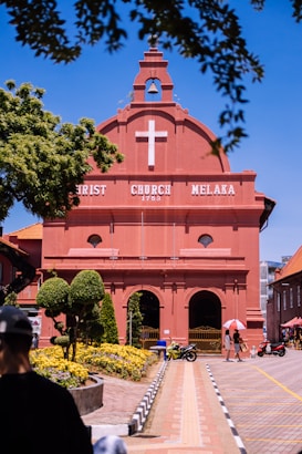 A red brick colonial-style church with a white cross, surrounded by well-maintained topiary and flower gardens. A clear blue sky frames the structure, with people walking nearby, some using umbrellas for shade. Motorbikes are parked on the pathway leading to the church entrance.