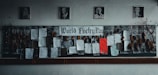 A bulletin board dedicated to World Poetry Day is adorned with various poems, some typed and others handwritten, pinned onto a dark fabric. Several framed portraits hang above the board. Dry leaves and twigs are arranged on each side of the board, adding an artistic touch.