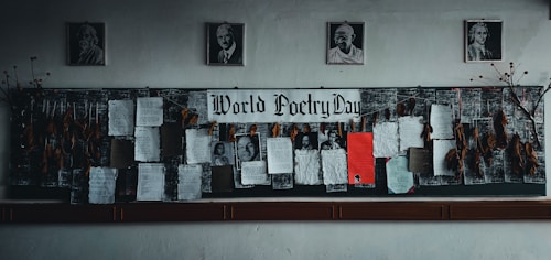 A bulletin board dedicated to World Poetry Day is adorned with various poems, some typed and others handwritten, pinned onto a dark fabric. Several framed portraits hang above the board. Dry leaves and twigs are arranged on each side of the board, adding an artistic touch.