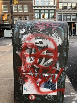 A mailbox covered in various graffiti and stickers is prominent in the foreground with an urban street scene in the background. The mailbox features layers of spray paint, including bold red lines and multiple tags overlaying each other. Behind it, there are brick buildings with windows, and a few traffic lights are visible, suggesting a city environment.
