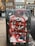 A mailbox covered in various graffiti and stickers is prominent in the foreground with an urban street scene in the background. The mailbox features layers of spray paint, including bold red lines and multiple tags overlaying each other. Behind it, there are brick buildings with windows, and a few traffic lights are visible, suggesting a city environment.