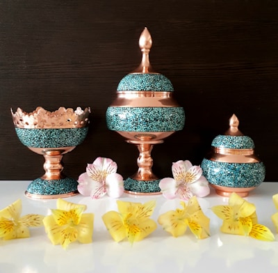 Three decorative containers made of turquoise and copper are artistically arranged. They feature intricate, mosaic-like patterns accented by striking copper tones. In front of them, a collection of vibrant yellow and pink flowers adds a natural contrast, set against a dark background and a reflective white surface.