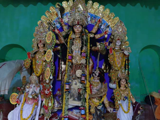 A colorful and vibrant depiction of several Hindu deities. The central figure is a multi-armed goddess adorned with intricate jewelry and a traditional headdress, holding various symbolic items. Surrounding her are other deities, each with distinct attributes and ornaments. The statues are placed against a green backdrop, and flower garlands are draped over the figures.