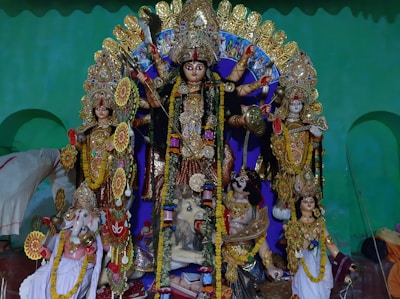 A colorful and vibrant depiction of several Hindu deities. The central figure is a multi-armed goddess adorned with intricate jewelry and a traditional headdress, holding various symbolic items. Surrounding her are other deities, each with distinct attributes and ornaments. The statues are placed against a green backdrop, and flower garlands are draped over the figures.