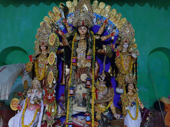 A colorful and vibrant depiction of several Hindu deities. The central figure is a multi-armed goddess adorned with intricate jewelry and a traditional headdress, holding various symbolic items. Surrounding her are other deities, each with distinct attributes and ornaments. The statues are placed against a green backdrop, and flower garlands are draped over the figures.