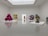 A modern art gallery featuring various sculptures and artworks in a spacious, minimalistic room. On the left, there is a large, reflective, magenta-colored sculpture. Near the center, a colorful geometric painting and a metallic orange sculpture are visible. On the right, a large, realistic blue bird sculpture is on display, with additional abstract shapes and a reflective ring artwork nearby. The floor and walls are white, enhancing the vibrant colors of the artworks.