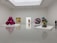 A modern art gallery featuring various sculptures and artworks in a spacious, minimalistic room. On the left, there is a large, reflective, magenta-colored sculpture. Near the center, a colorful geometric painting and a metallic orange sculpture are visible. On the right, a large, realistic blue bird sculpture is on display, with additional abstract shapes and a reflective ring artwork nearby. The floor and walls are white, enhancing the vibrant colors of the artworks.