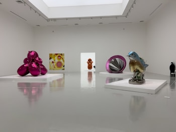 A modern art gallery featuring various sculptures and artworks in a spacious, minimalistic room. On the left, there is a large, reflective, magenta-colored sculpture. Near the center, a colorful geometric painting and a metallic orange sculpture are visible. On the right, a large, realistic blue bird sculpture is on display, with additional abstract shapes and a reflective ring artwork nearby. The floor and walls are white, enhancing the vibrant colors of the artworks.