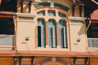 An architectural facade featuring elegant arched windows with shuttered panels set against a beige wall. Decorative tiles with a green and turquoise geometric pattern border the windows. Two outdoor lights are mounted on the wall, casting distinct shadows. The structure exhibits elements of colonial-style architecture with a tiled roof and an intricate wooden overhang.
